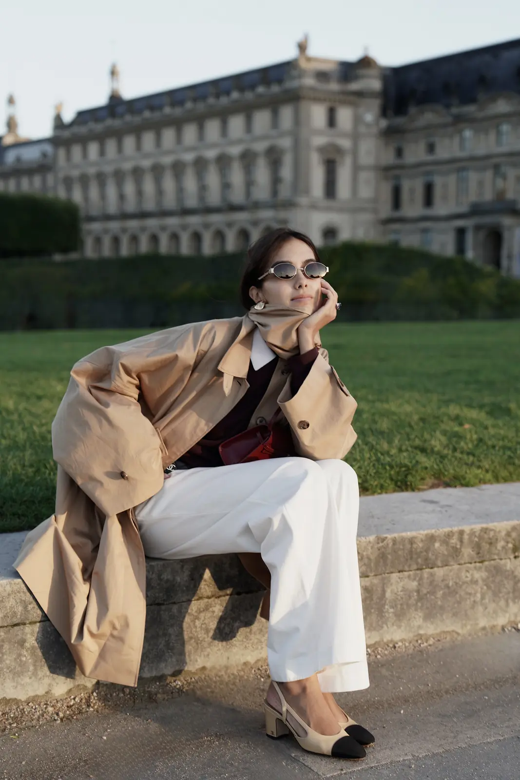 Sharon seated outdoors in a beige trench coat, white trousers, and sunglasses.
