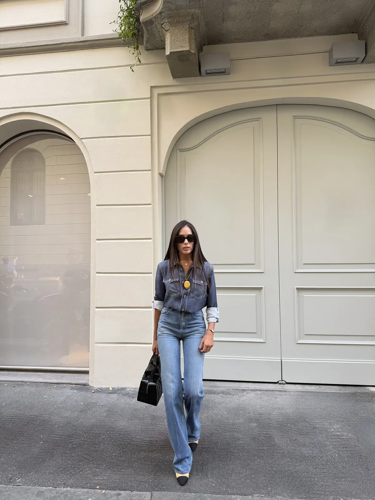 Sharon in a denim shirt, blue jeans and two-tone shoes, carrying a black bag outside a doorway in Milan.