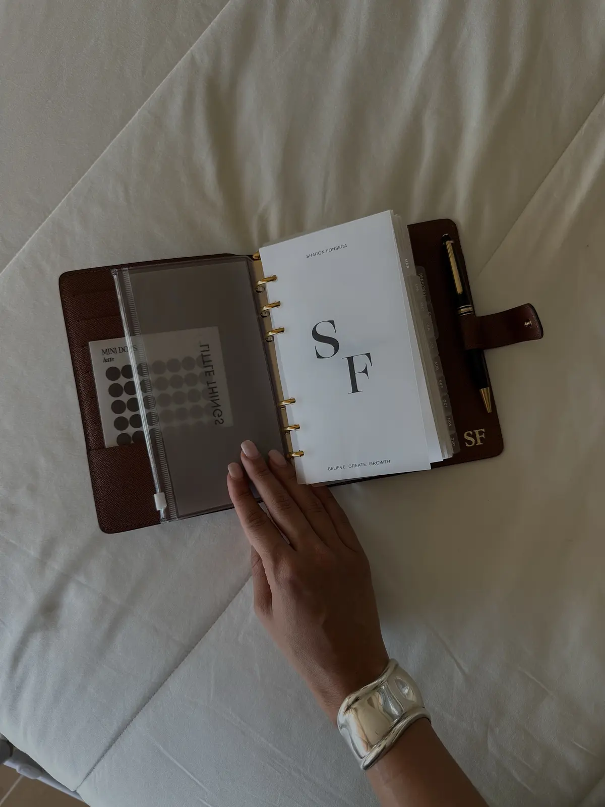 An open brown planner with Sharon Fonseca’s SF monogram, a pen and a hand resting on the pages.