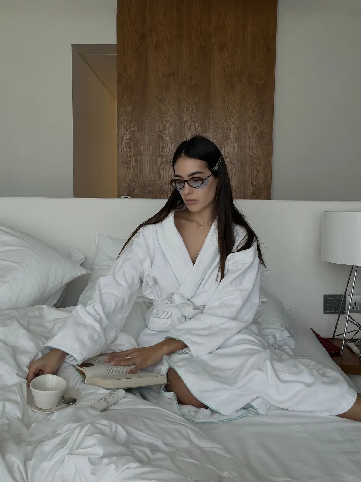 A woman in a white robe reads on a bed beside a cup.